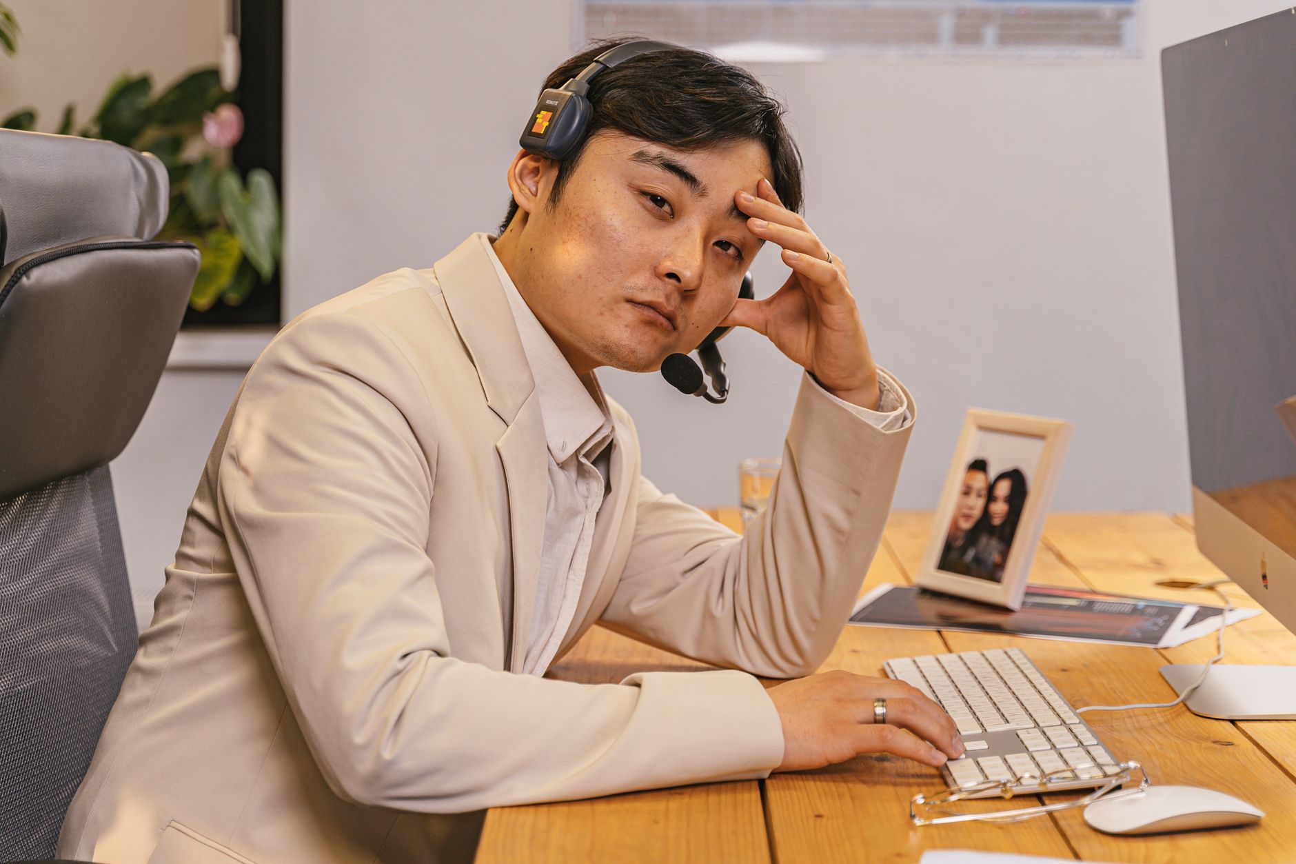 Asian man in beige suit working at computer with headset indoors.