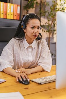 Asian woman using computer and headset in a modern office setting.