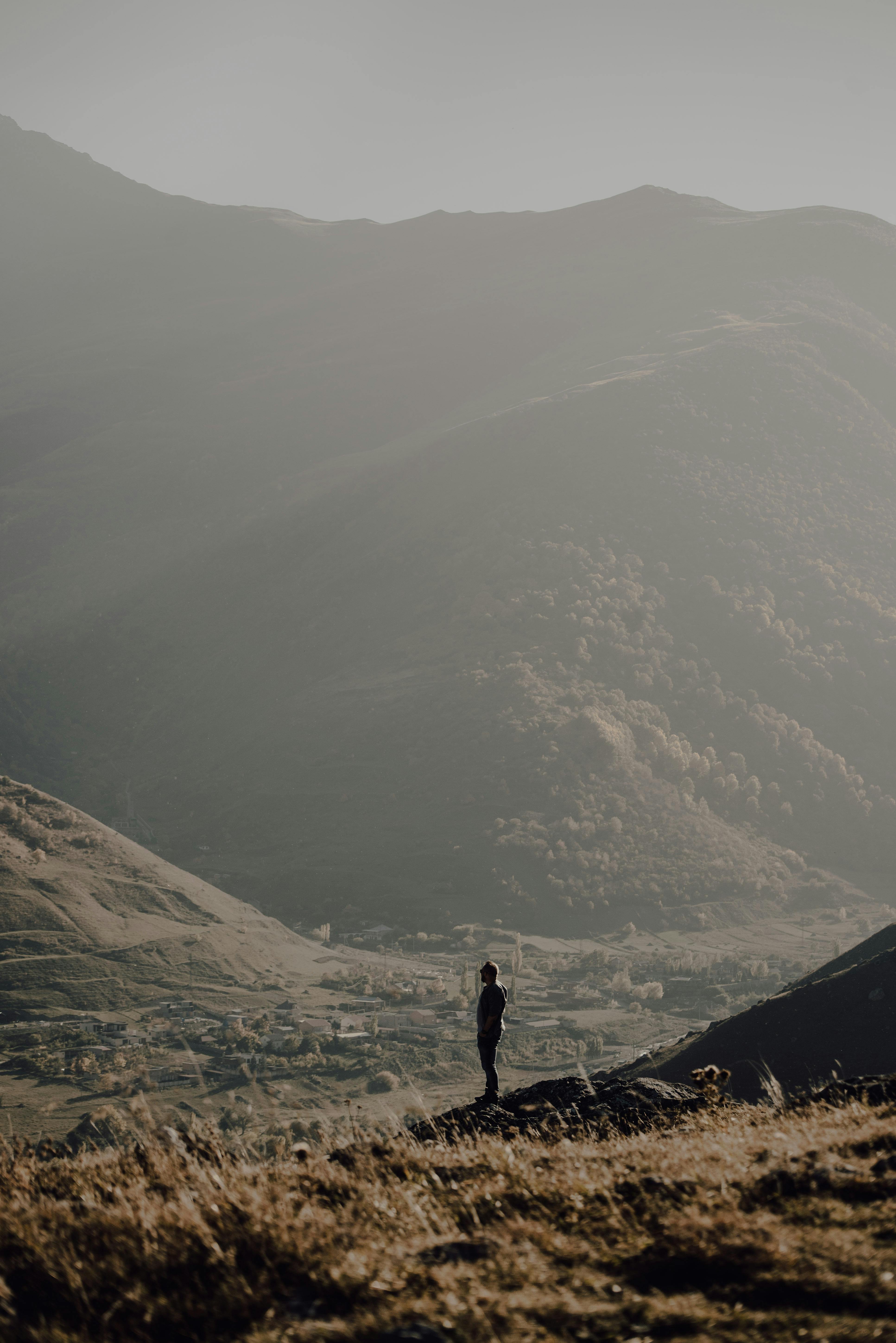 A lone man stands amidst vast mountains, under a soft, muted sky.
