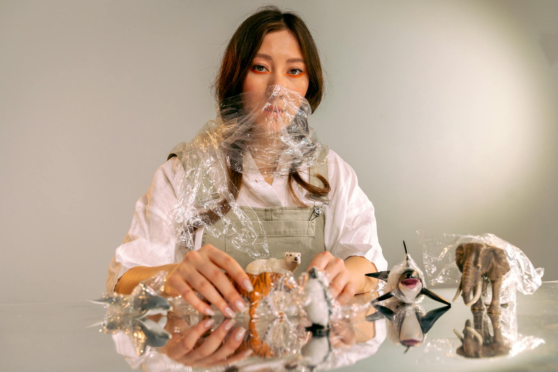 Asian woman artistically posing with plastic-wrapped toy animals, hinting at environmental themes.