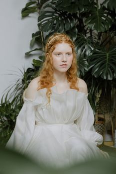 A young redhead woman in a flowing white dress sits gracefully indoors, surrounded by lush green foliage.