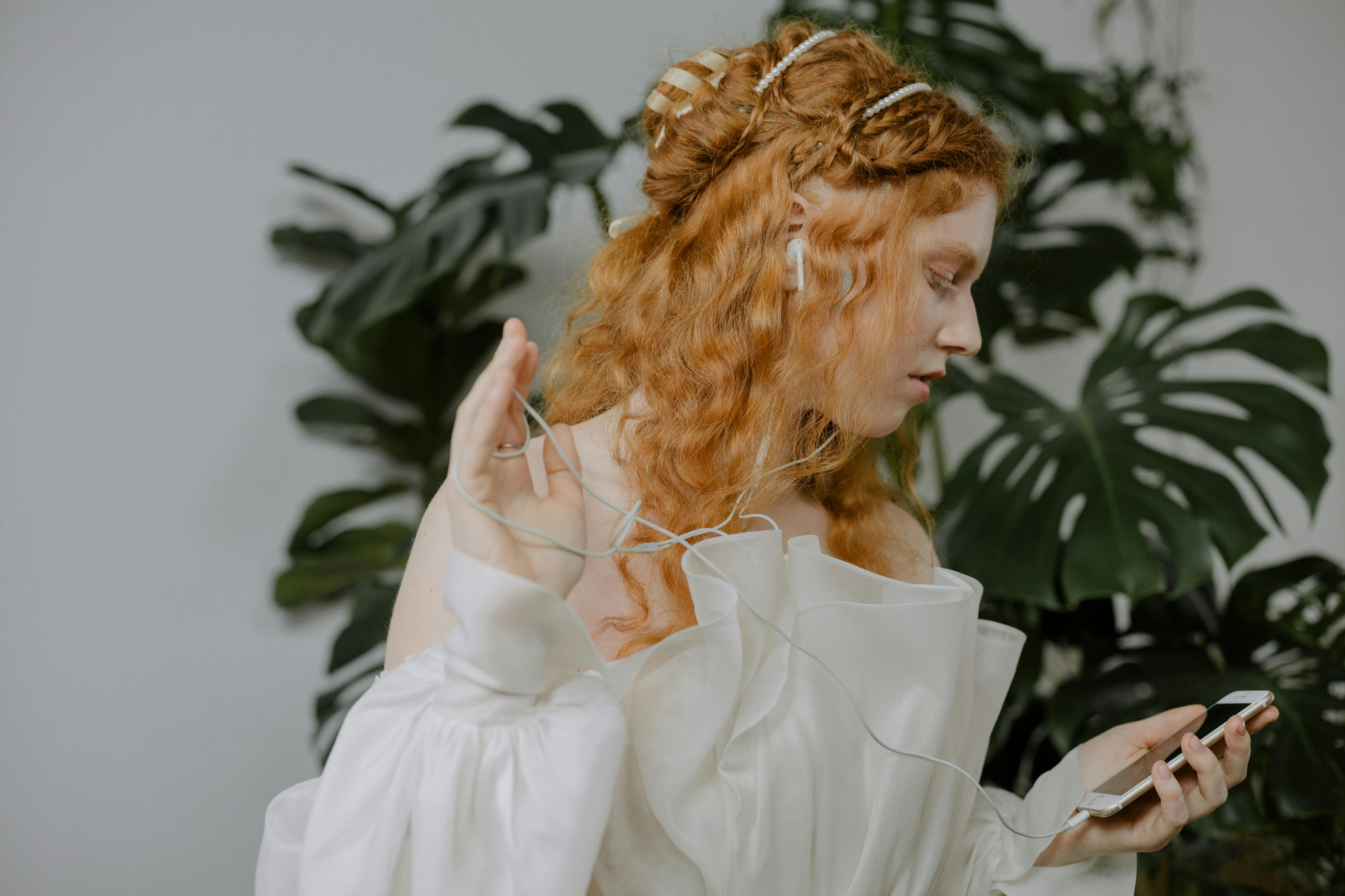 Redheaded woman in white dress listening to music on smartphone with earphones, indoors with lush plants.