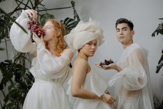 A stylish studio portrait of three individuals in white attire holding grapes, surrounded by green leaves.