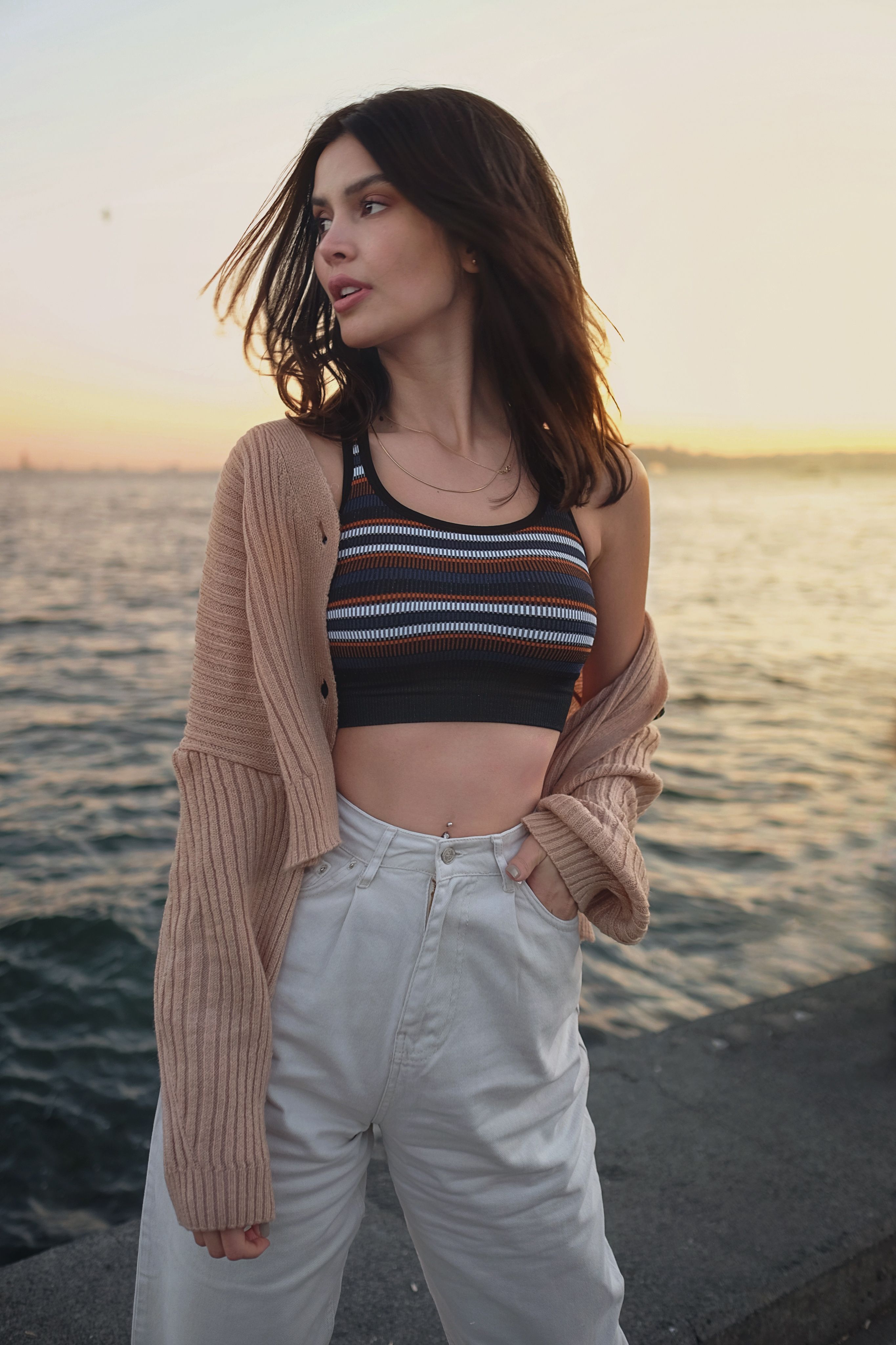 Calm female with hand in pocket in white trousers looking away near wavy sea washing coast in sunset