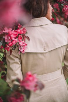 Back view of unrecognizable female in stylish coat strolling near lush apple tree with small tender blooming flowers in garden