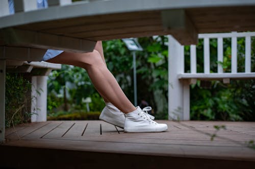 Free Crop anonymous female in stylish white sneakers sitting on wooden bench in park with lush green bushes on summer day Stock Photo