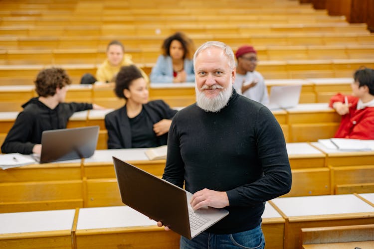 Professor In Black Turtleneck Sweater Holding Silver Macbook In Classroom