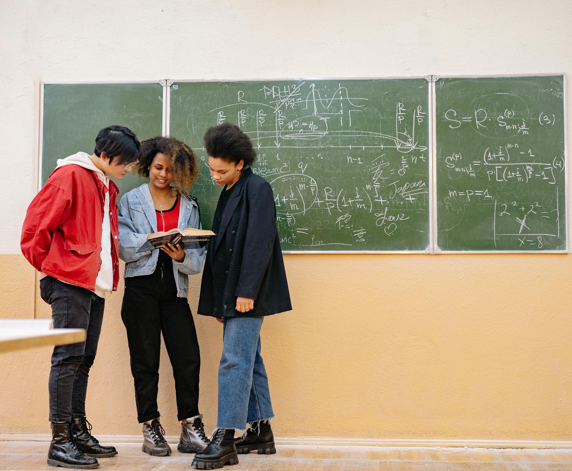 Three diverse students standing by a chalkboard with complex equations, collaborating on a math problem.