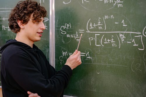 Young Man Standing next to a Blackboard and Pointing at a Math Equation 