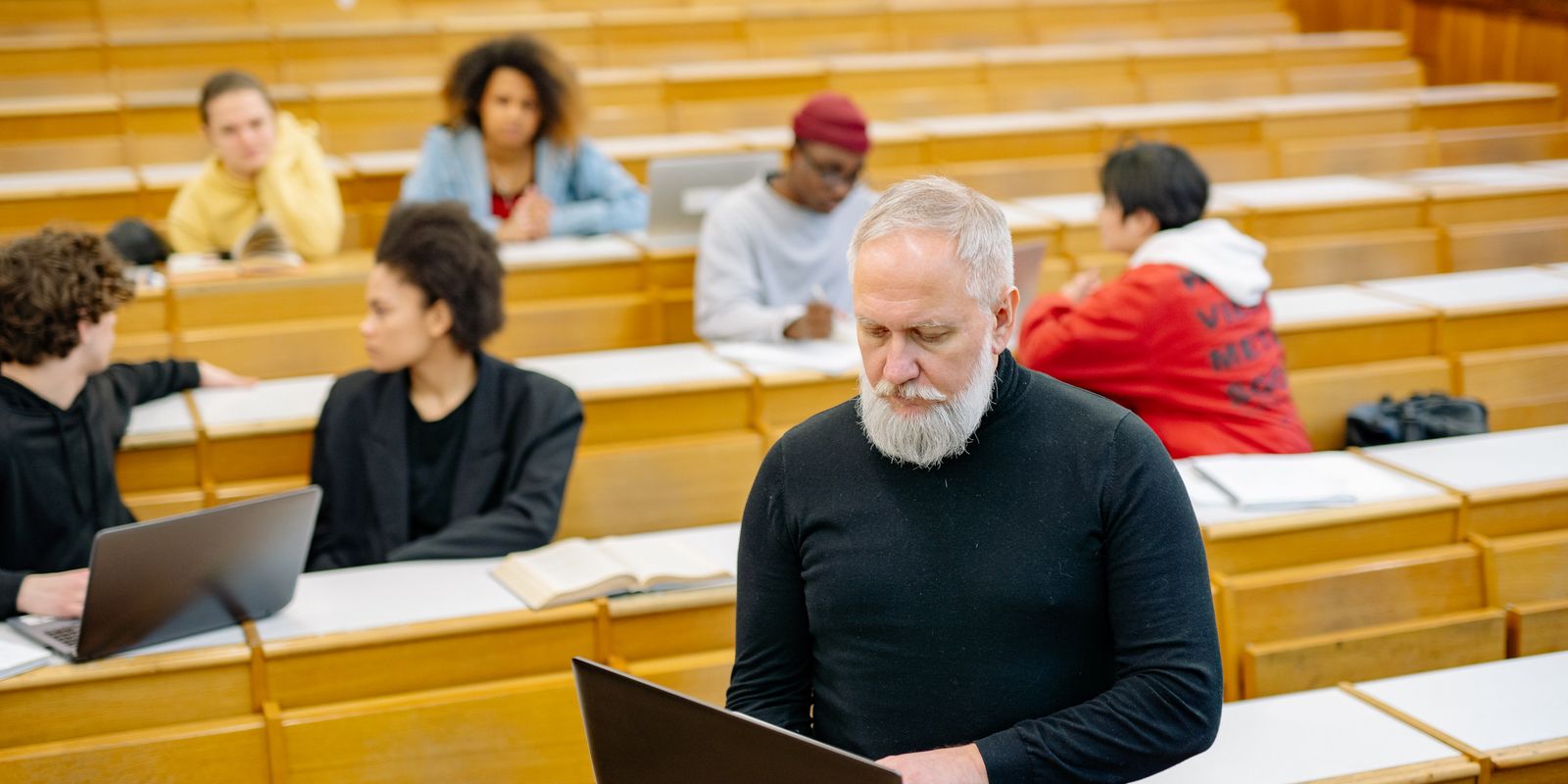 Mature teacher interacting with diverse students using laptops in a spacious lecture hall setting.