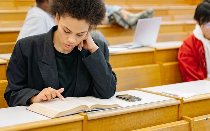 A young woman focuses intently on reading a book in a college lecture hall.