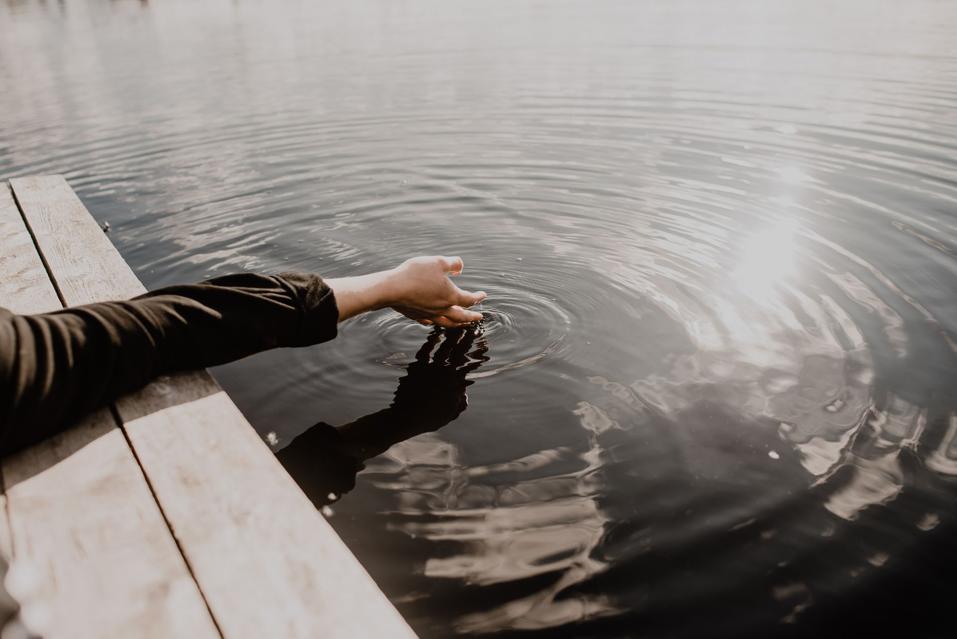 A serene scene of a hand gently touching the calm water surface, creating ripples in the sunlight.