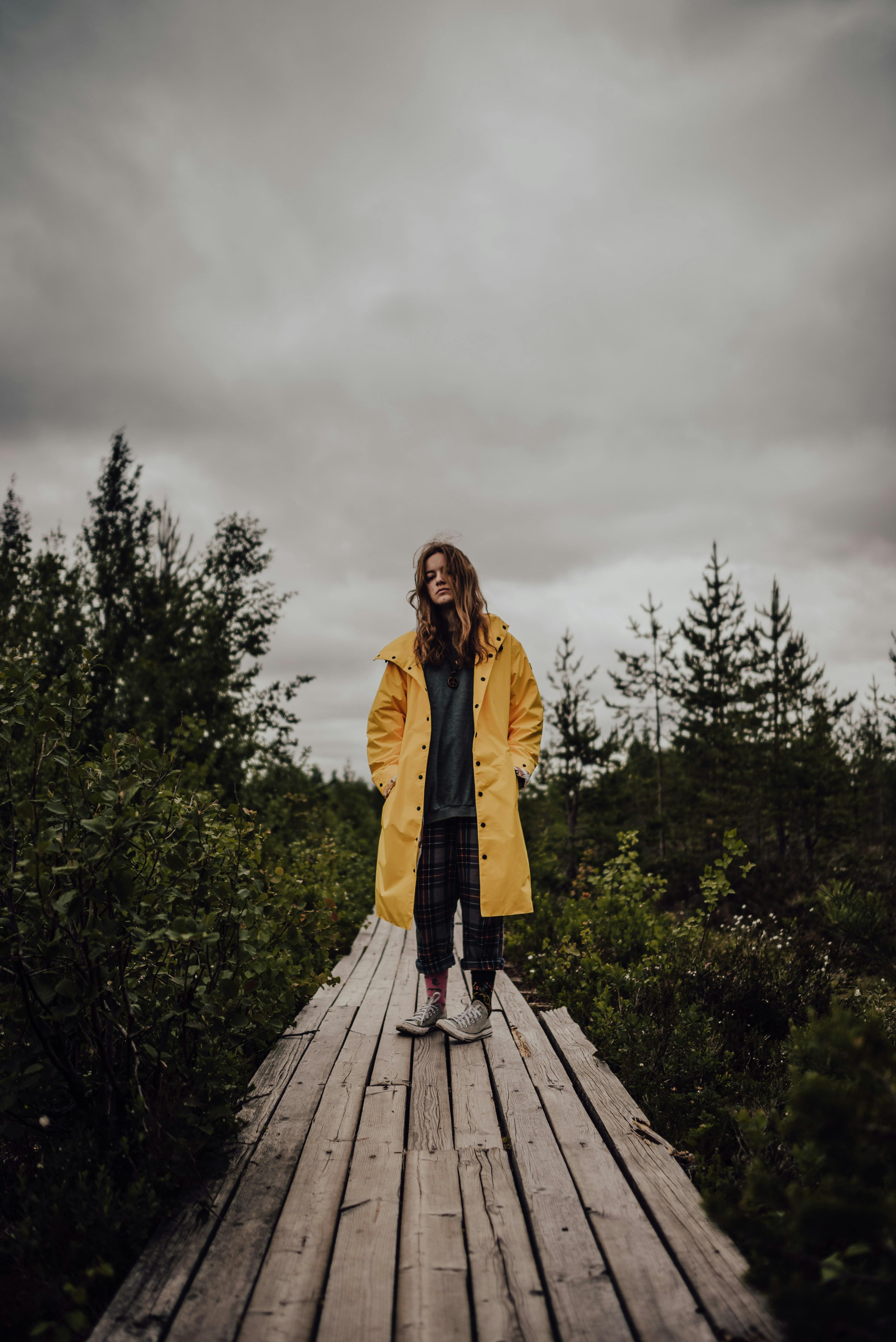 A woman in a yellow coat stands on a wooden walkway amidst lush greenery, under a cloudy sky.