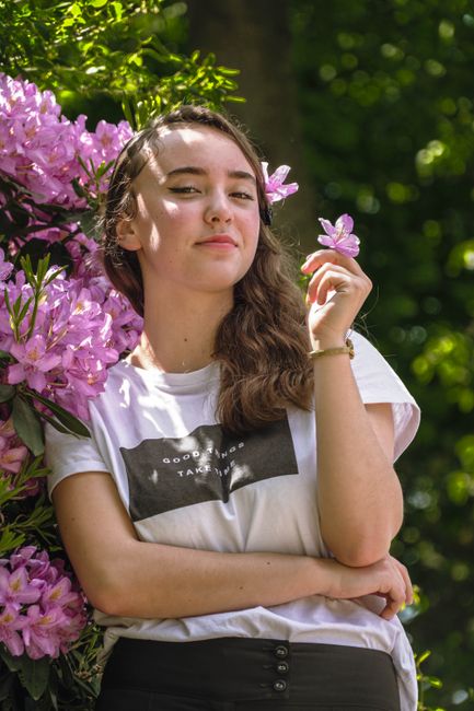 Portrait of a young woman posing confidently with pink flowers in a sunny outdoor garden.