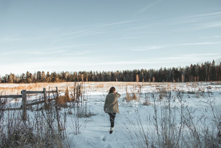 A woman walks through a snowy field surrounded by trees on a clear winter day.