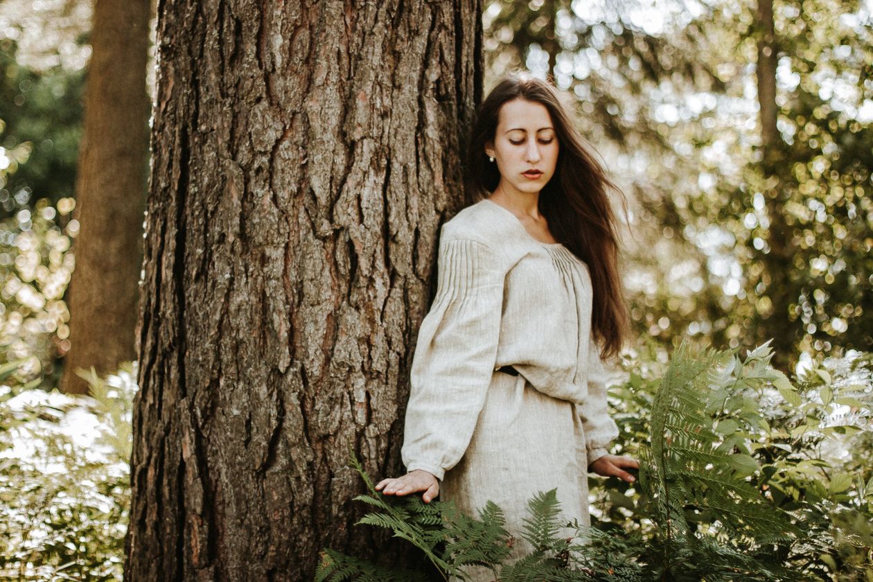 Woman wearing a natural linen long-sleeve dress beside a tree in a forest