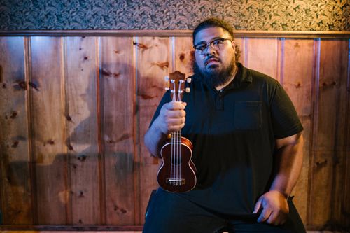 Free A bearded man sitting indoors holding a ukulele, wearing a black polo shirt. Stock Photo