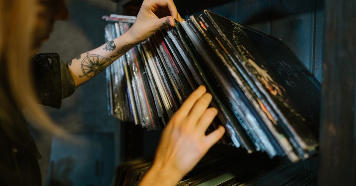 A woman with tattoos browses vinyl records on a shelf in a music studio setting.