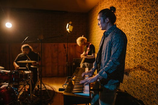 A band rehearsing in a warmly lit studio, featuring drums, keyboard, and electric guitar.