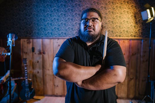Bearded drummer in a recording studio, holding drumsticks with crossed arms.