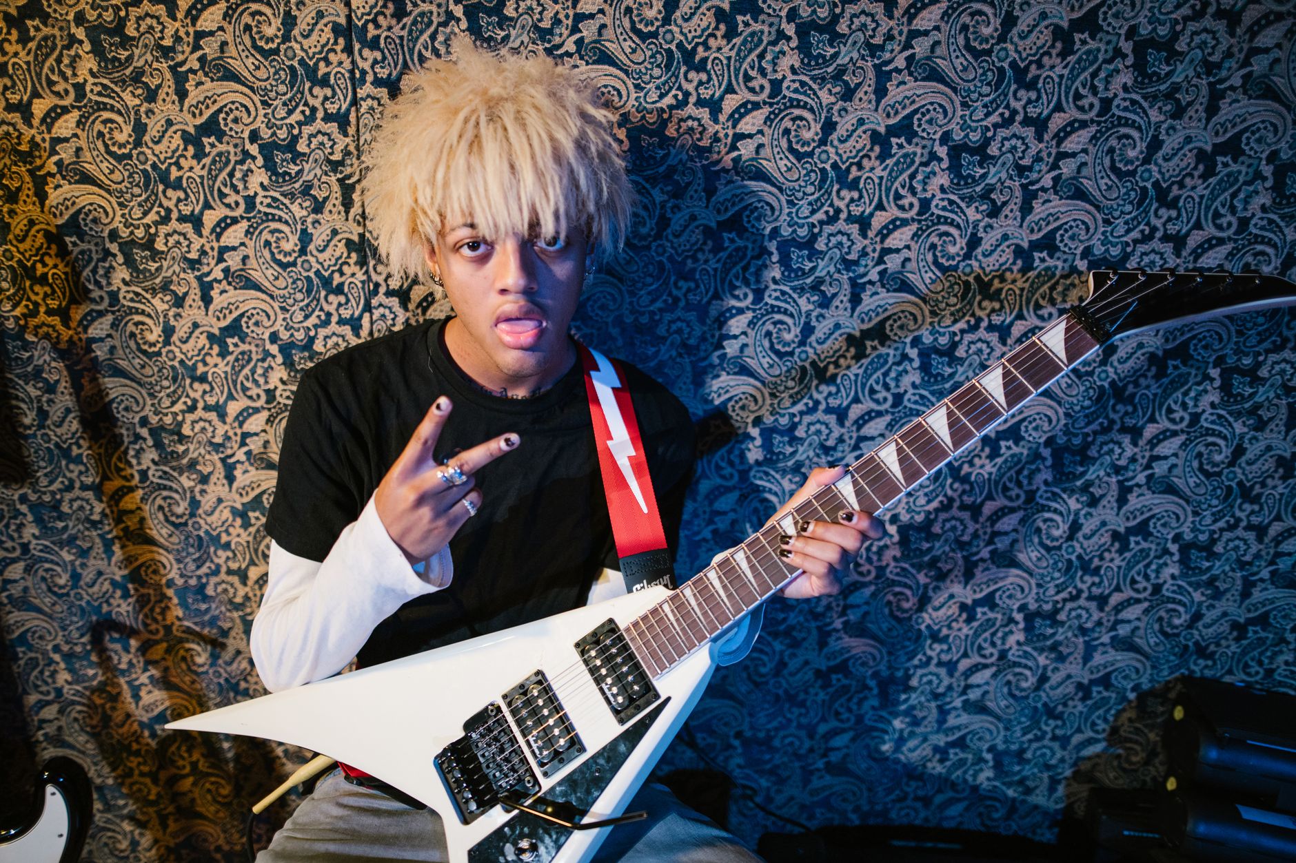A young musician poses confidently with an electric guitar in an indoor setting featuring a patterned background.