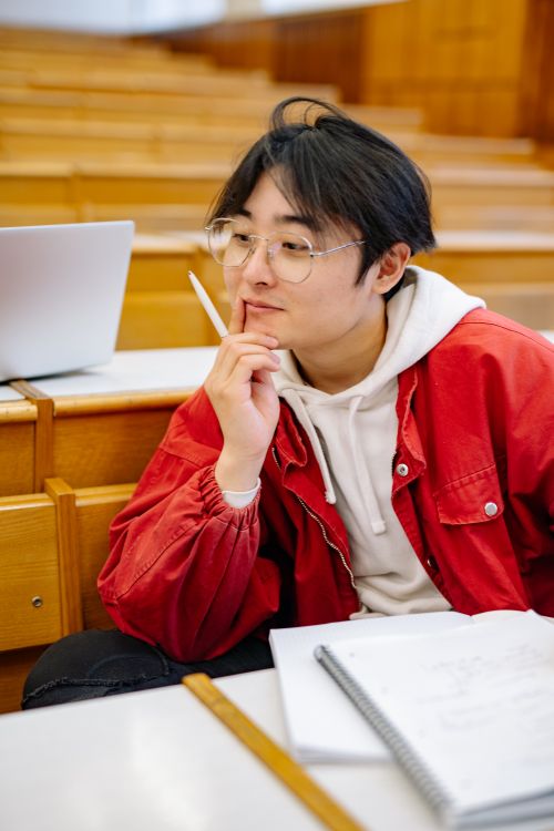 Free Asian male student in red jacket thinking during class in lecture hall. Stock Photo