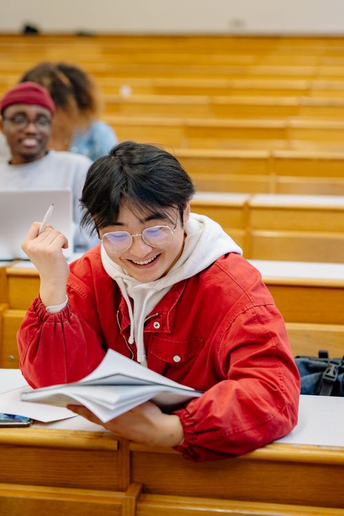 Free A smiling student in a classroom setting, wearing a red jacket, reading and taking notes. Stock Photo