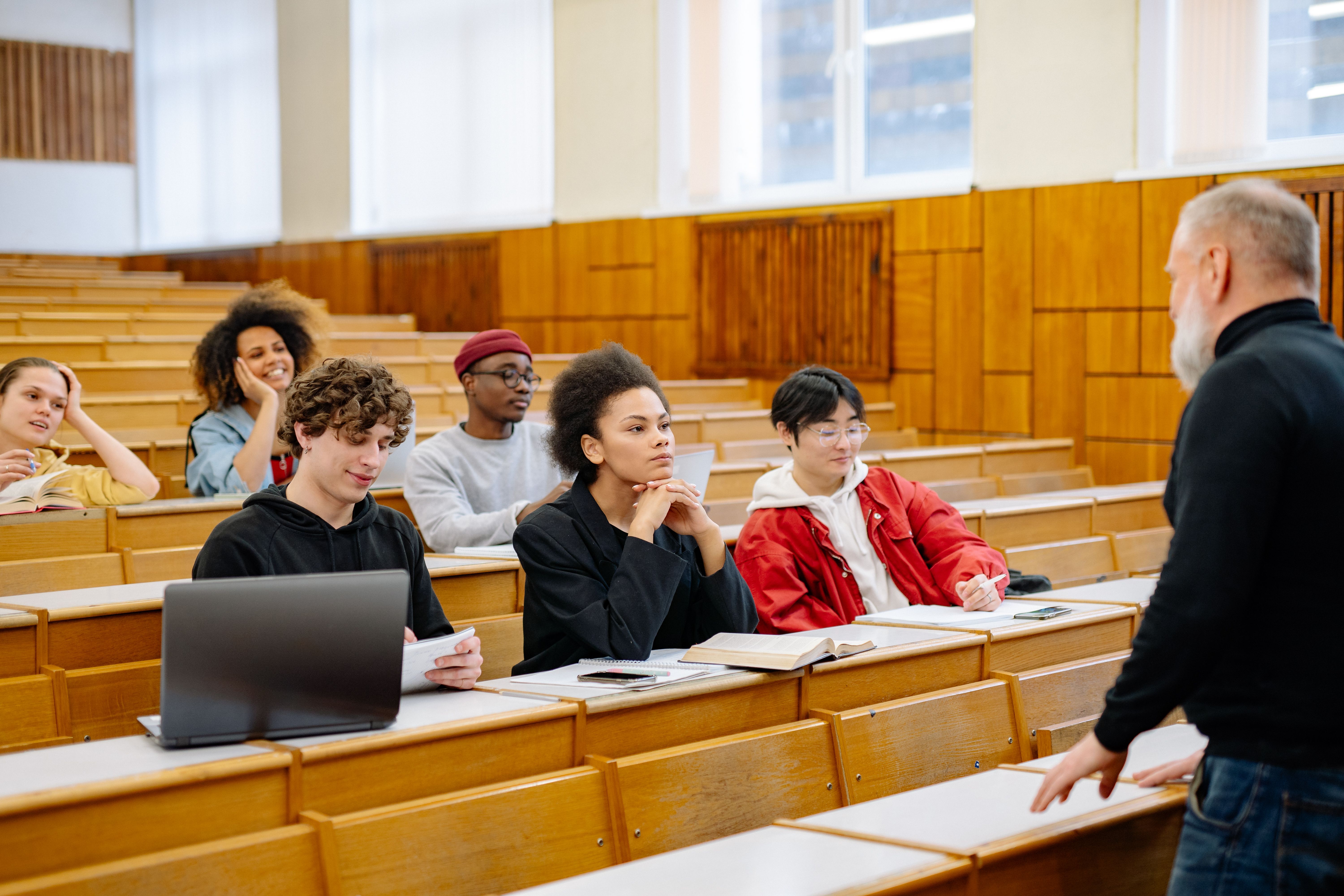 Students seated in a lecture hall listening to a professor, with laptops and books on desks