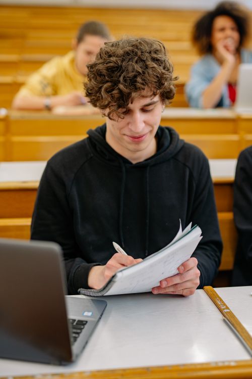 Free A focused student in a black hoodie writing notes during a classroom lecture. Stock Photo