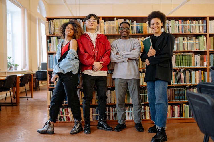 Friends Standing Near The Bookshelves In The Library