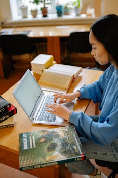 A young woman is focused on studying with her laptop at a wooden desk with open books.