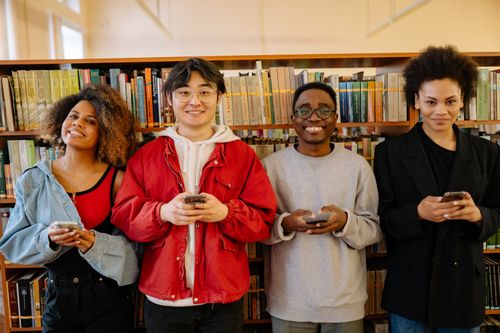 Free Cheerful group of diverse students using smartphones in a library environment, smiling and engaged. Stock Photo