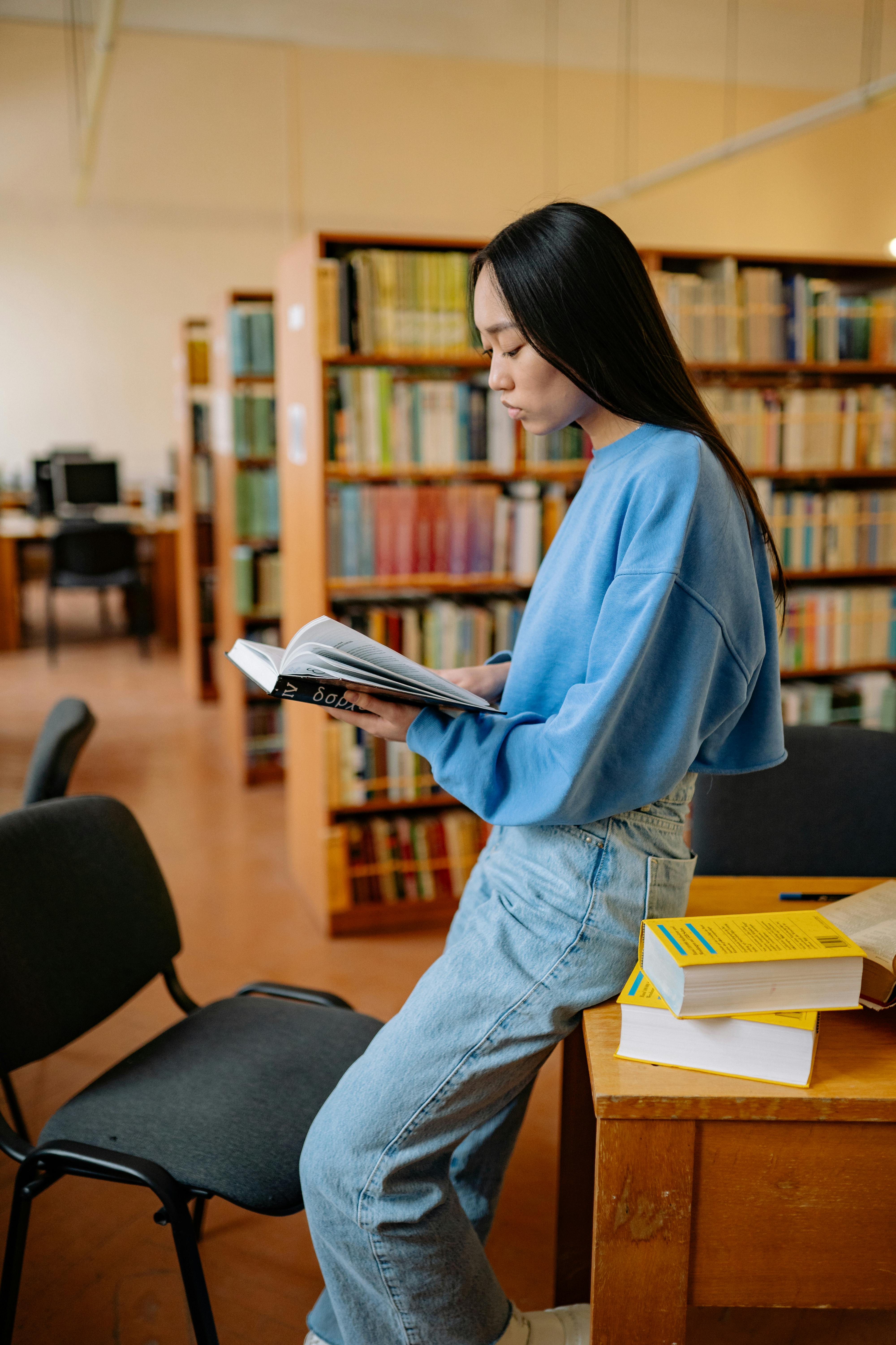 Free A young woman attentively reading a book in the library, surrounded by shelves filled with books. Stock Photo
