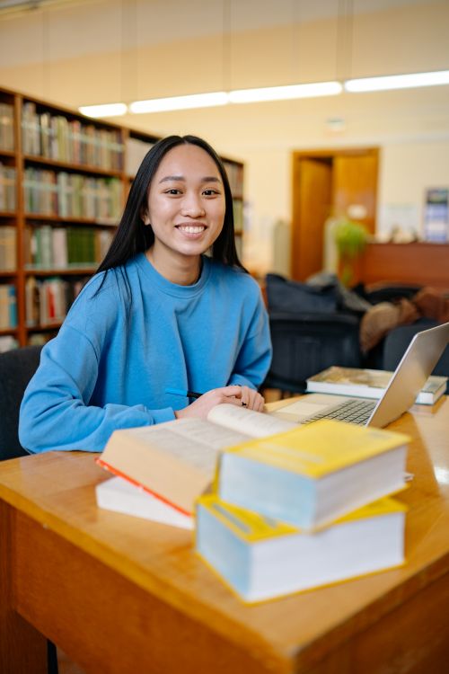 Free A young woman student sitting in a library with books and a laptop, smiling while studying. Stock Photo