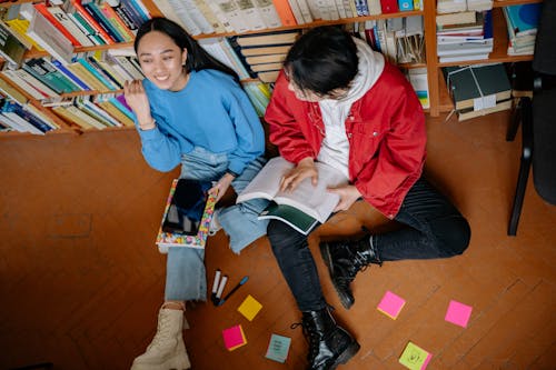 High-Angle Shot of Man and Woman Sitting on the Floor while Leaning on Book Shelf