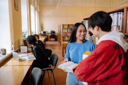 Free Two smiling university students talking and holding books in a library setting. Stock Photo
