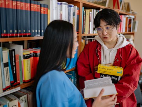 Two students conversing in a university library surrounded by bookshelves.