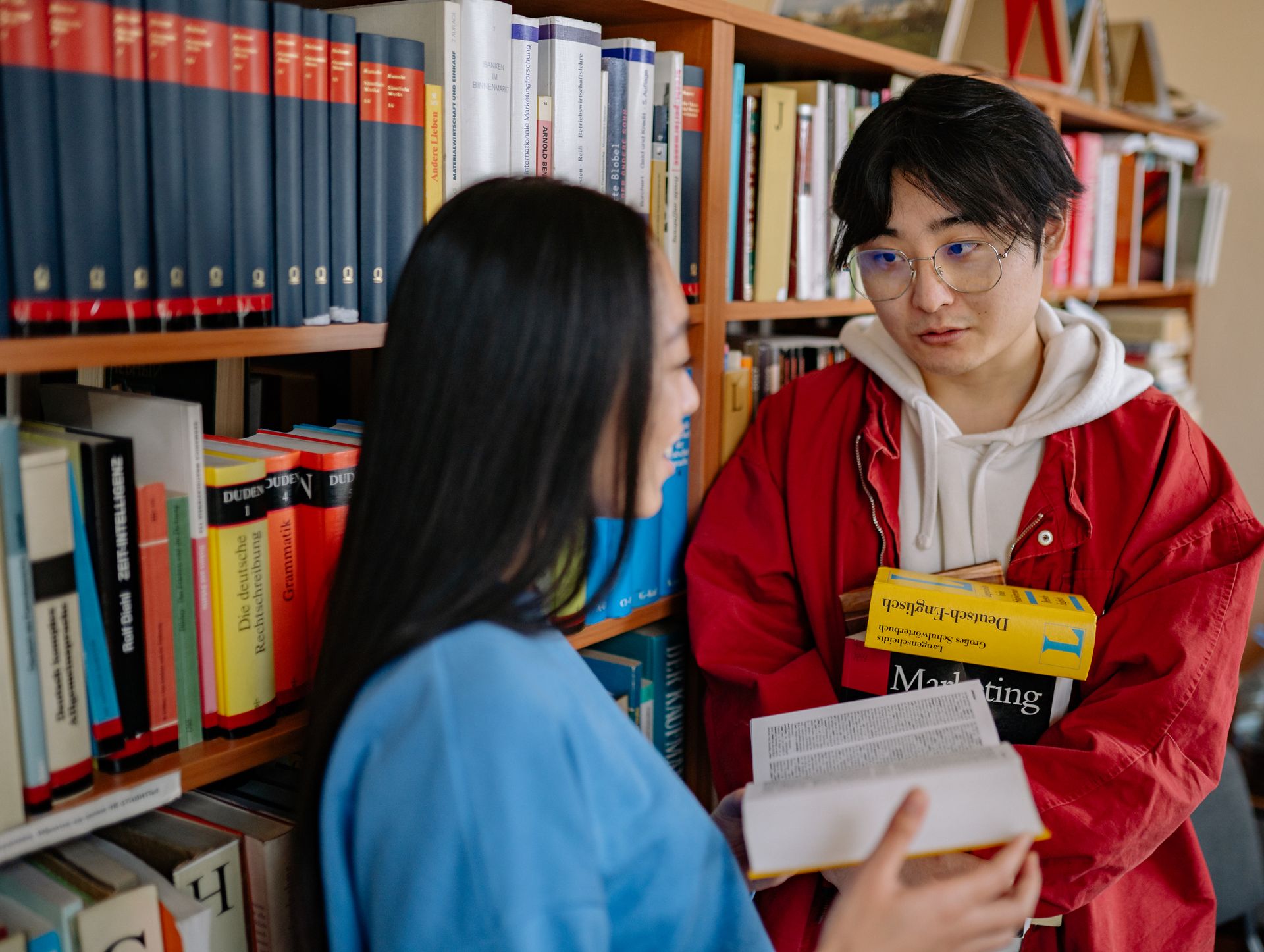 Two students conversing in a university library surrounded by bookshelves.