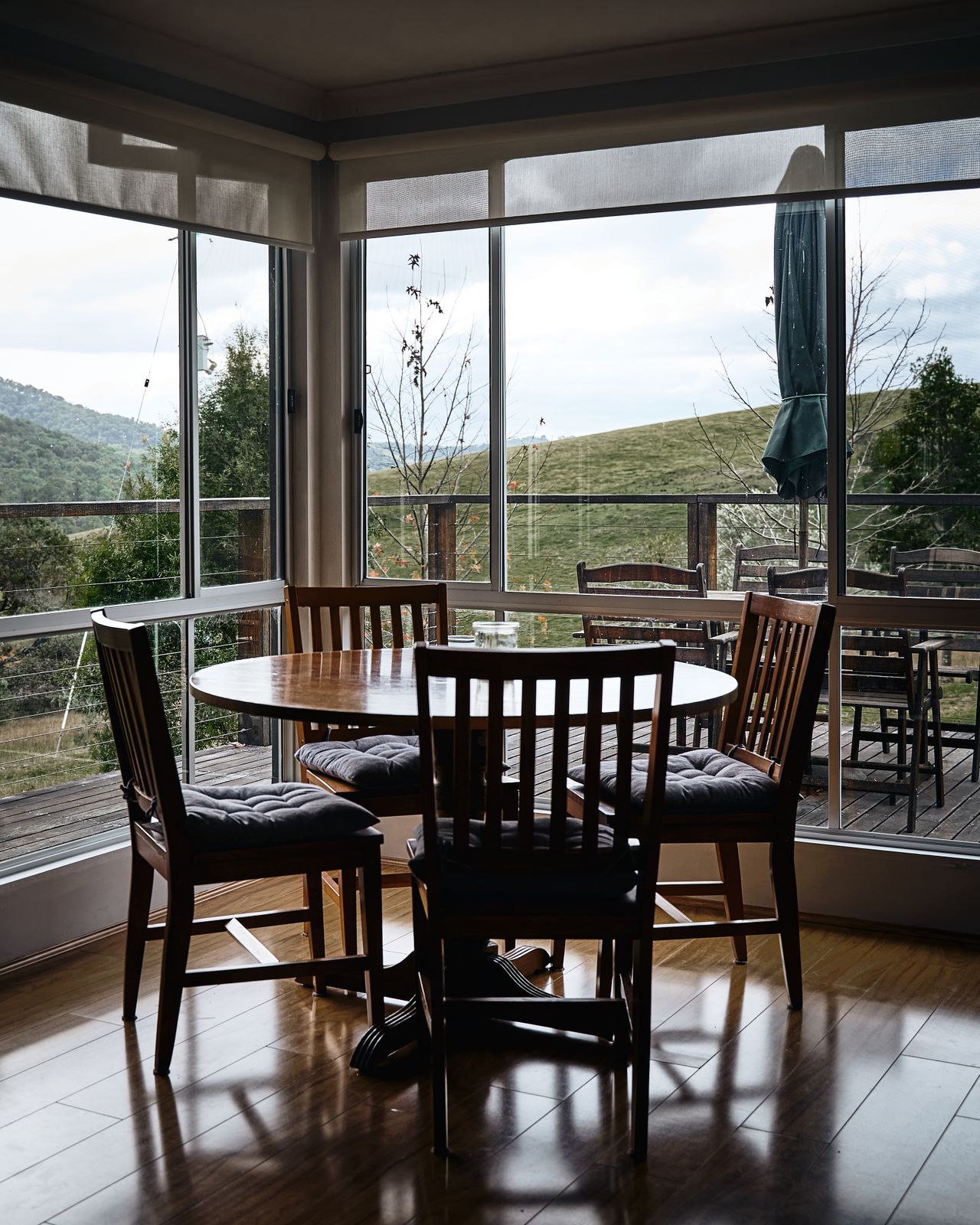 Round wooden dining table with four chairs beside large windows overlooking hills