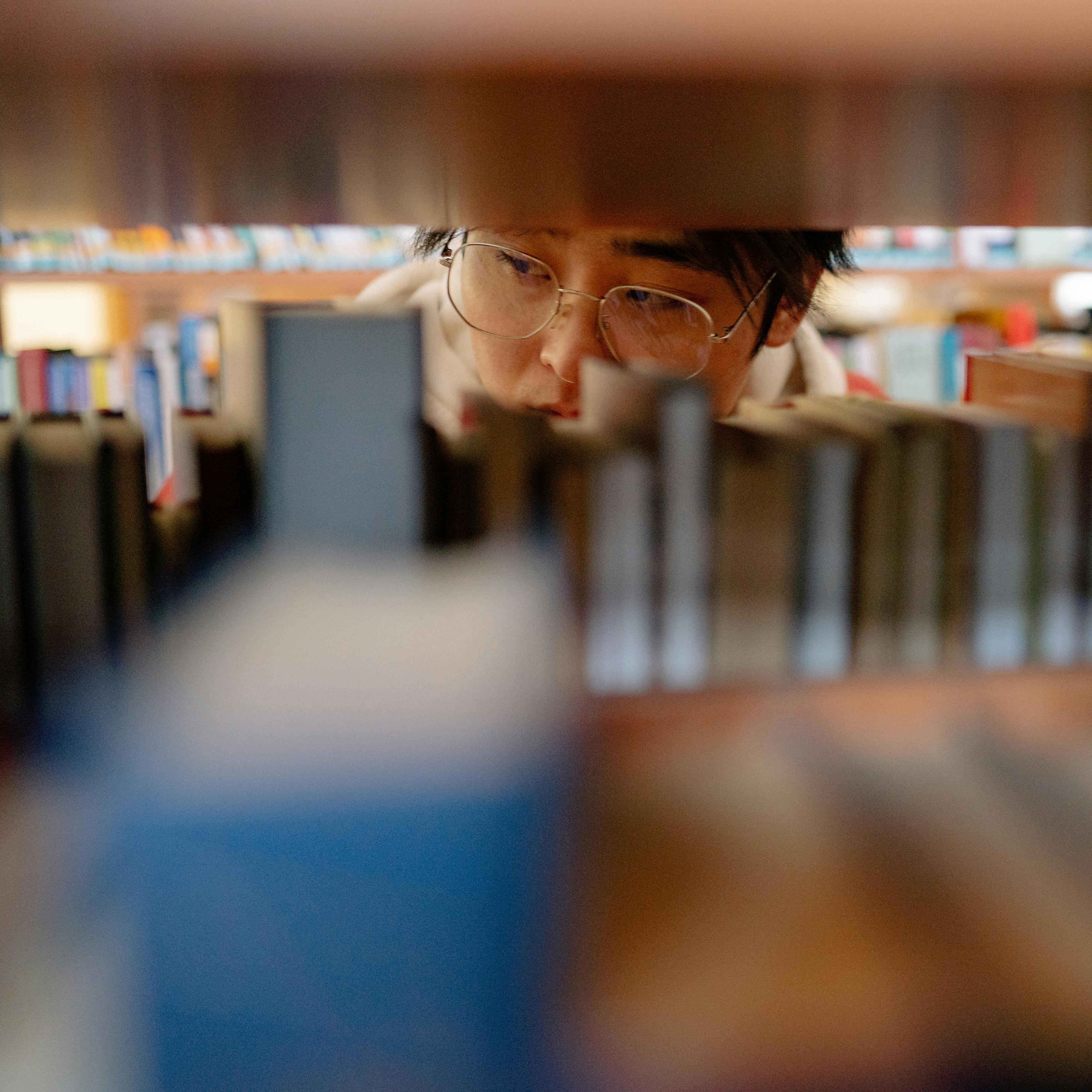 Asian man with eyeglasses looking at books on a library shelf