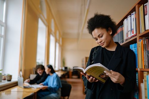 Free A young woman reads a book in a serene library environment, showcasing a focused academic atmosphere. Stock Photo