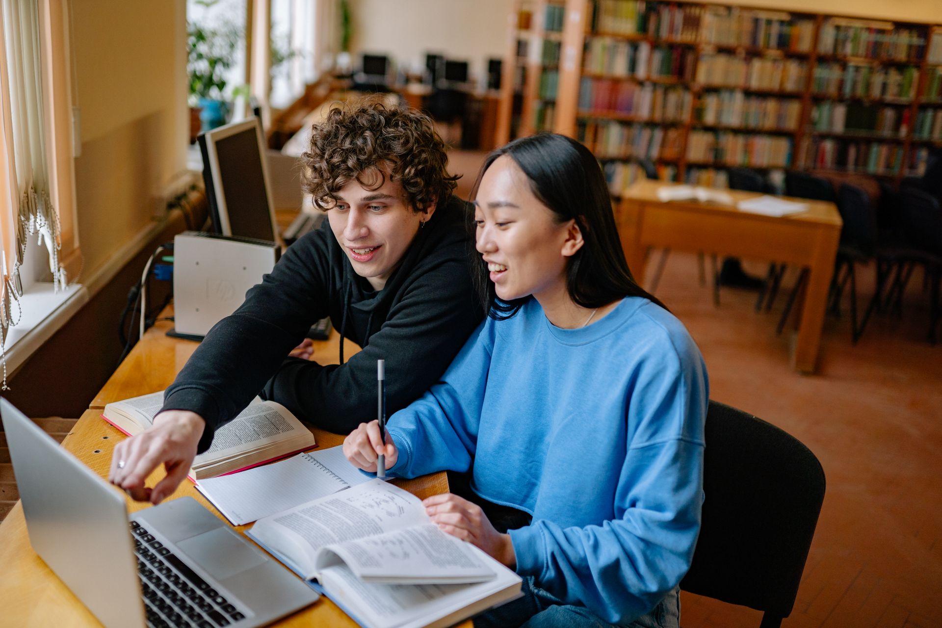 Two students studying together in a library with books and a laptop.