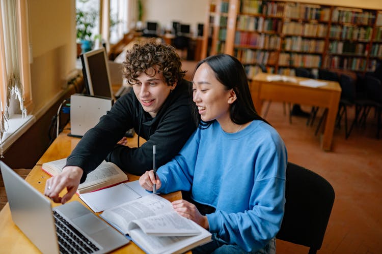 Students Studying In A Library Using Laptop And Books 