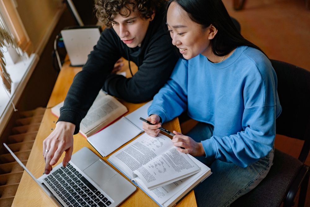 Two students learning side by side at a laptop with a caring guide nearby