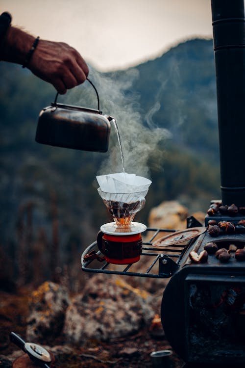 Unrecognizable male hand of traveler pouring hot water from portable metal kettle in cup making coffee while resting in campsite in mountains
