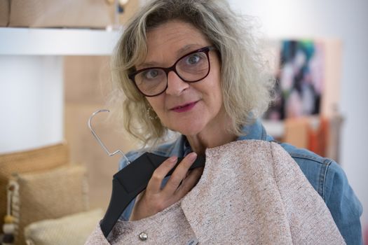Elderly woman with glasses shopping and smiling while choosing stylish clothing.