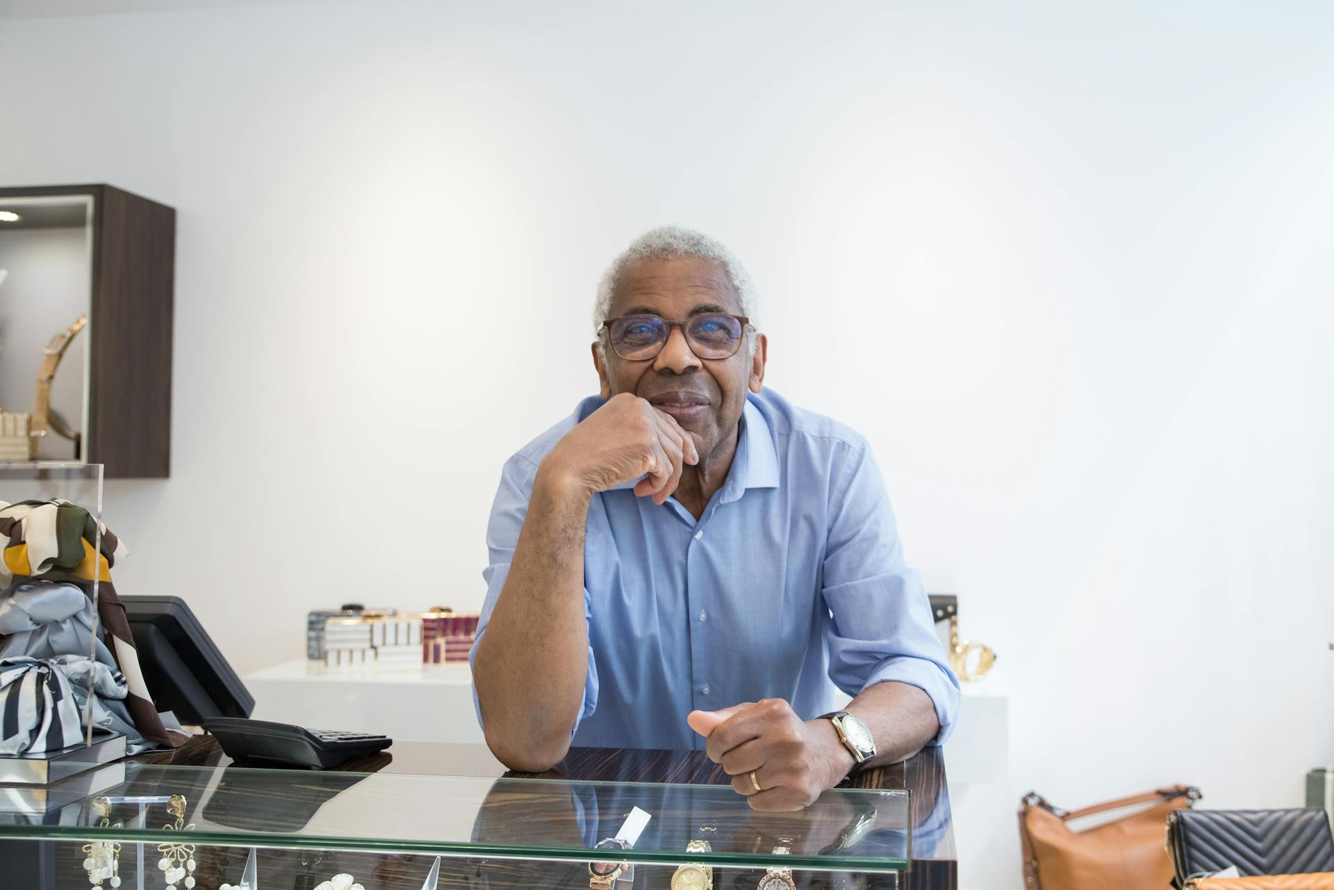 Elderly man with glasses leaning on a counter, smiling in a retail store setting.