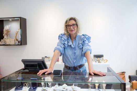 A senior woman stands confidently at a jewelry store counter in Portugal, showcasing elegance.
