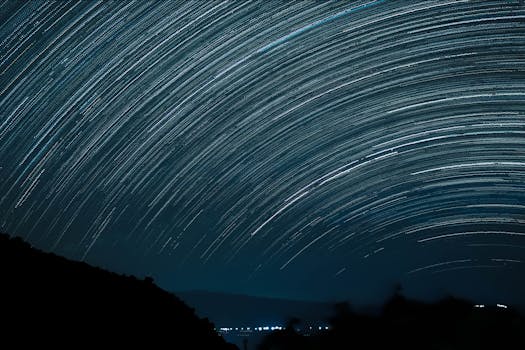 Dramatic long exposure of star trails arching over a mountainous landscape, showcasing the beauty of space at night.