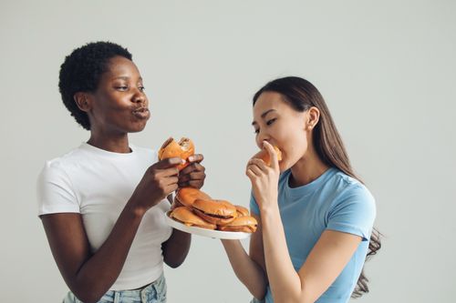 Free Two women sharing a moment eating burgers indoors, highlighting enjoyment and camaraderie. Stock Photo